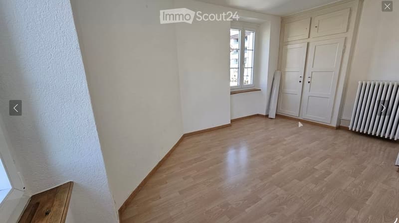 Empty room with wooden flooring, white walls, and a window with a small sill. There are two white doors and a radiator on the right side.
