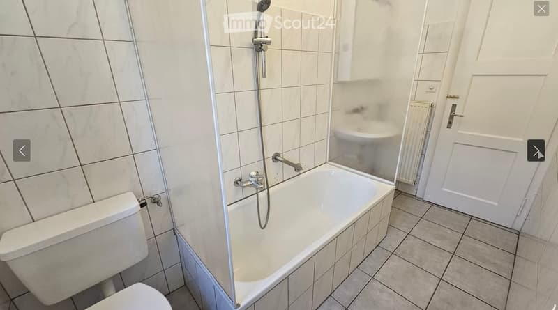 Modern bathroom with a white bathtub, toilet, and showerhead. The walls are tiled in a light, neutral color, and the floor is tiled with a similar tone. A glass shower enclosure is visible, and a door leads to another room.