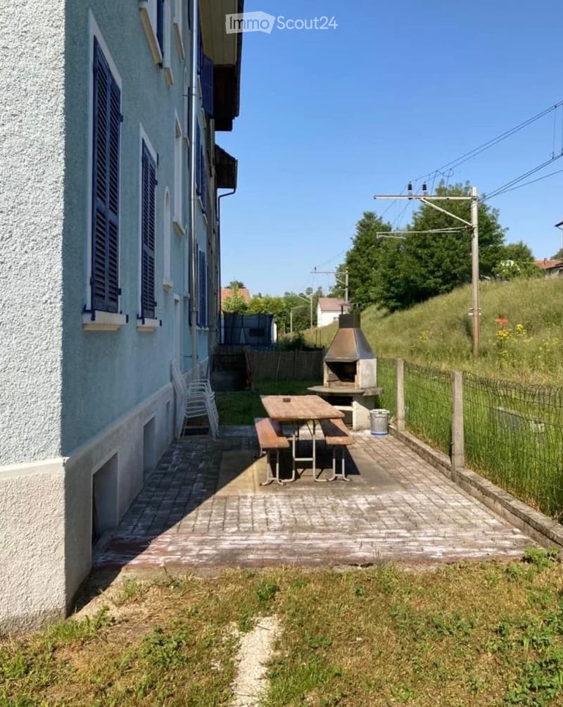 Outdoor patio area with a brick floor, a wooden picnic table, benches, a barbecue, and a stack of white chairs against the building.