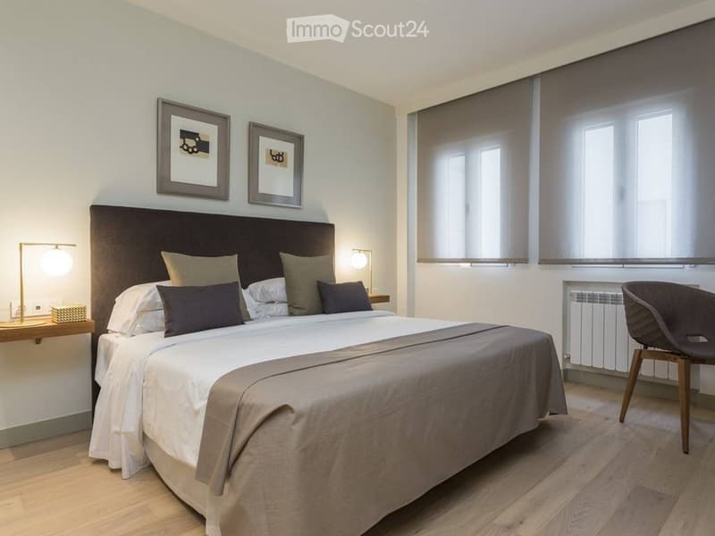 Modern bedroom with a large bed, nightstands, lamps, a chair, and framed artwork on the wall.