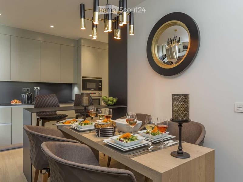 Elegant dining area with a wooden table set for six, surrounded by modern chairs, wine glasses, and plates of food. A stylish lamp and a round mirror adorn the walls, with a sleek kitchen area visible in the background.