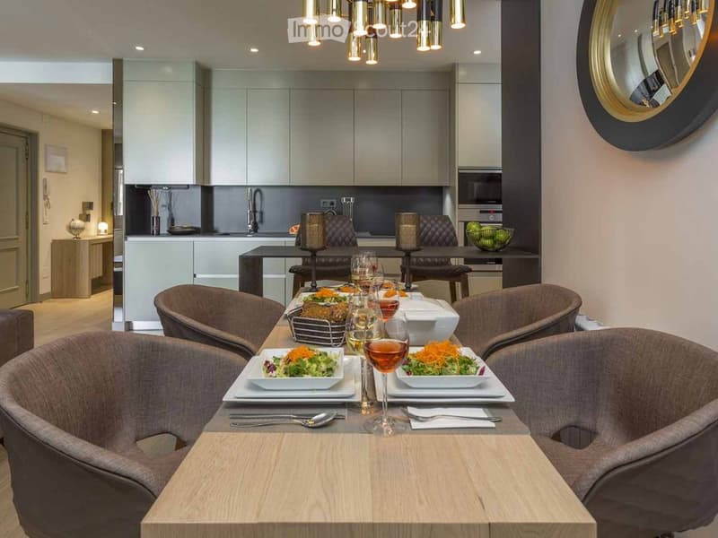 Elegant dining area with a wooden table set for a meal, surrounded by modern chairs, with a kitchen area in the background featuring sleek cabinets and appliances.