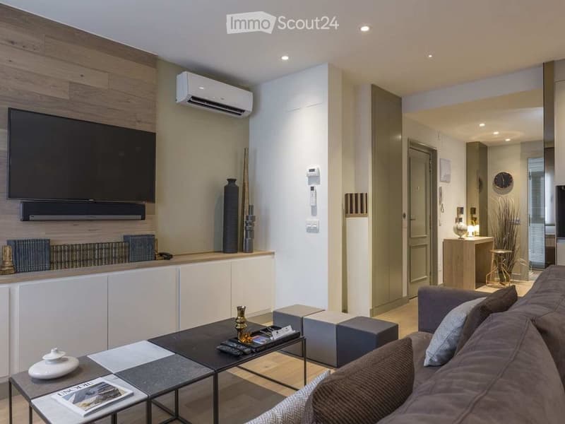 Modern living room with a grey sofa, coffee table, decorative vases, and a television mounted on a wooden wall. The space features built-in cabinets, a radiator, and a sleek design with warm lighting.