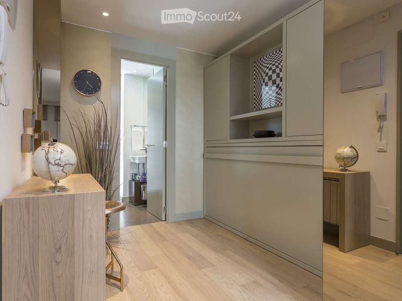 Modern hallway with light wooden flooring, a small wooden stool, a globe on a table, a wall-mounted clock, and an open doorway leading to a bathroom with a sink and mirror.
