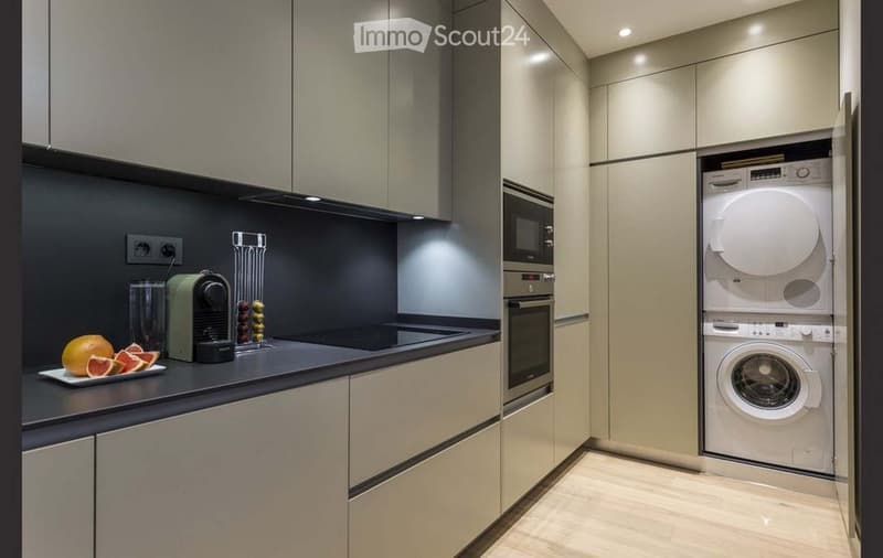 Modern kitchen with built-in oven, microwave, washing machine, and dishwasher. Countertop features a coffee maker, fruits, and a cup.