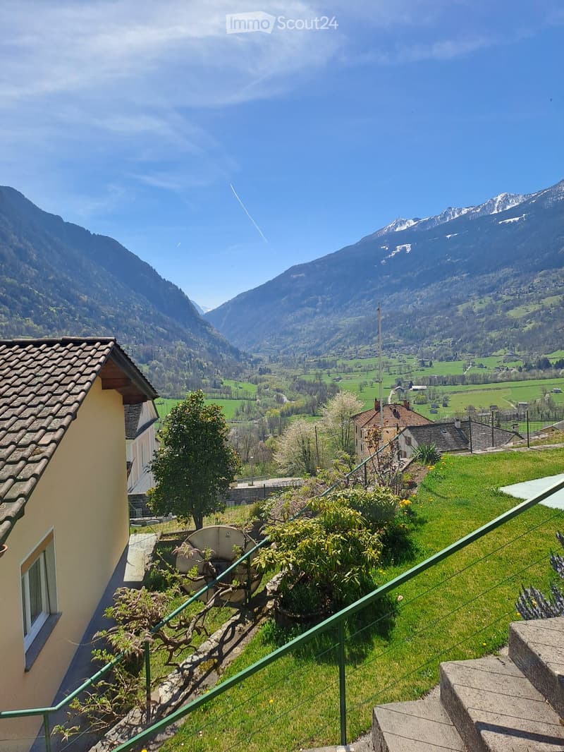 House with tiled roof, surrounded by a green lawn, steps leading to the entrance, distant view of mountains and a valley