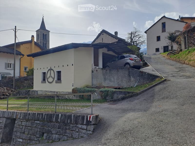 House with a car parked in the garage, located on a sloped road, surrounded by other houses and a church tower in the background.