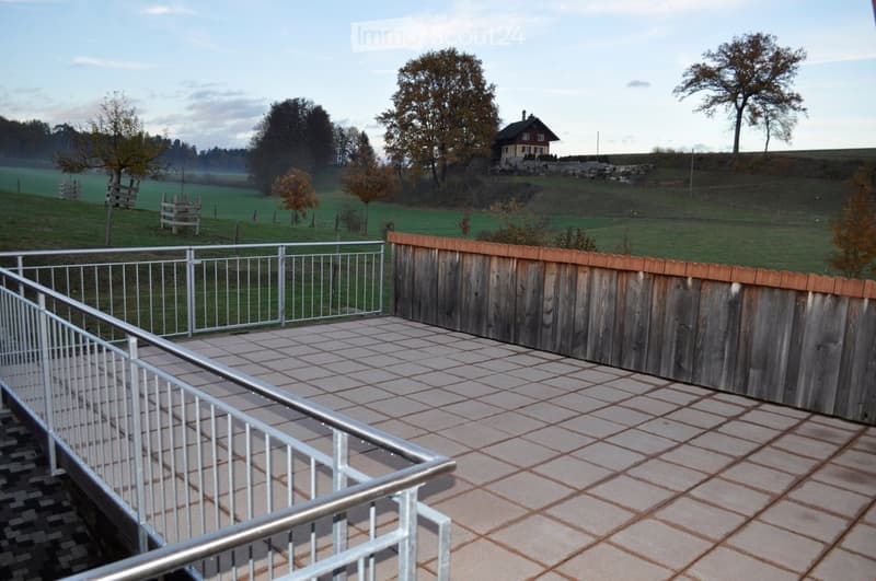 Balcony with tiled floor, metal railing, wooden fence, overlooking a grassy field with trees and a small house in the distance.