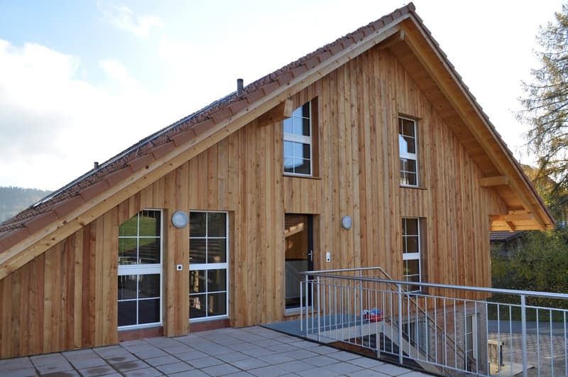 A modern wooden house with a sloped roof, large windows, and a raised ground floor accessed by a staircase.