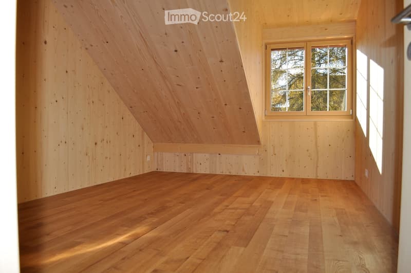 Empty attic room with wooden walls and floor, large window with view of trees, natural lighting.