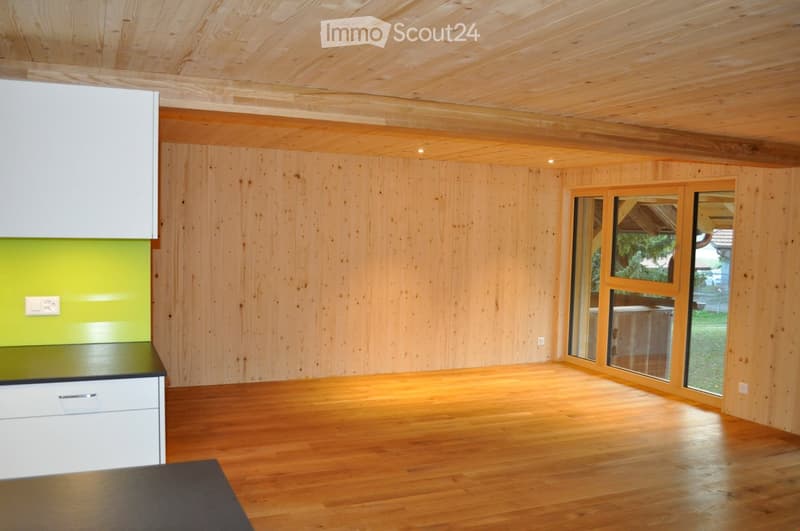 Empty room with wooden walls, ceiling, and floor, featuring a white cabinet and a green accent wall. Large windows provide natural light and a view of the outdoors.