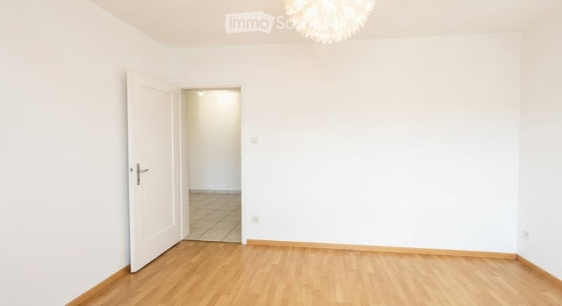 Empty room with light wooden flooring, white walls, and a chandelier hanging from the ceiling. An open door leads to another room with tiled flooring.