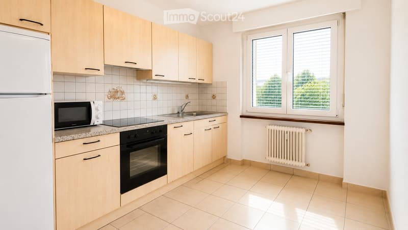 Modern kitchen with light wooden cabinets, granite countertop, stainless steel sink, microwave, oven, and radiator under the window.