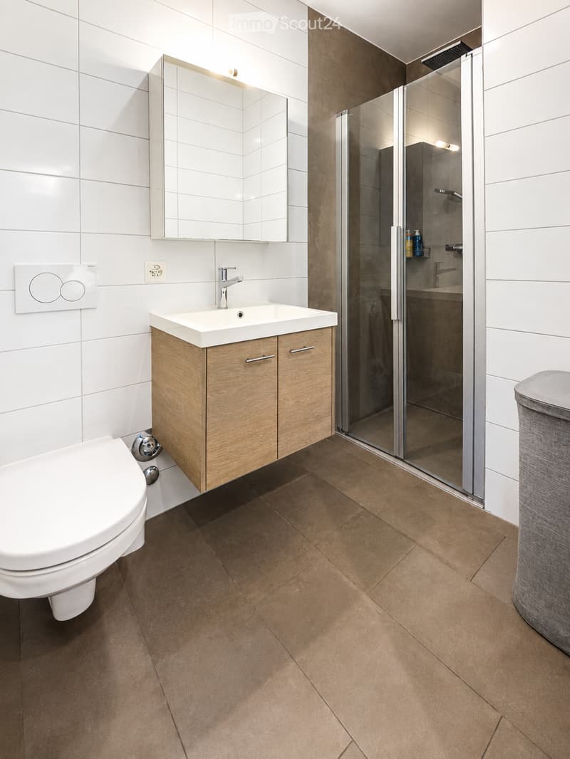 Modern bathroom with white tiles, a wooden sink cabinet, a toilet, a large mirror, and a glass-enclosed shower area.