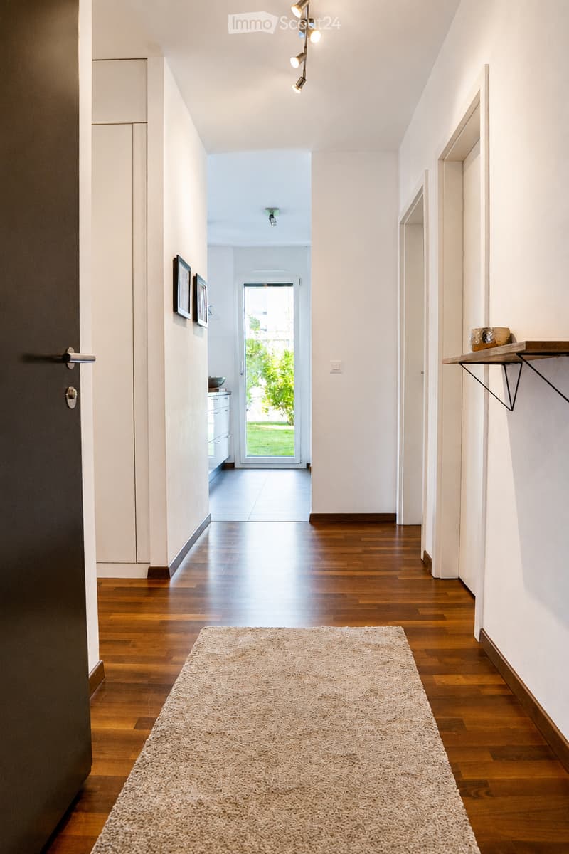 Modern hallway with wooden flooring, white walls, recessed lighting, and a small shelf with decorative items. A door leads to a bright room with a view of greenery.