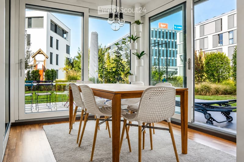 Bright dining area with a wooden dining table, white chairs, and a light-colored rug, overlooking a green outdoor space with modern buildings.