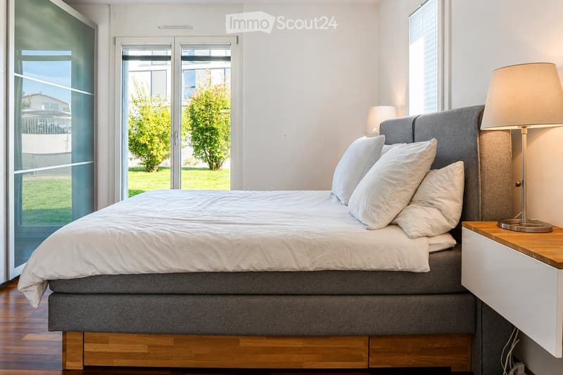 Modern bedroom with a large bed, white bedding, nightstand, and lamp. Large windows and sliding glass doors lead to a green outdoor area.
