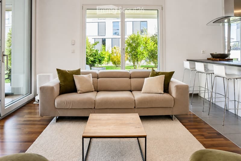 Modern living room with a beige sofa, wooden coffee table, white bar counter with stools, and large windows overlooking a green garden.