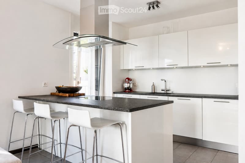 Modern kitchen with white cabinets, black granite countertop, and a central island with three white bar stools. Features a stand mixer, a bowl on the counter, and a sleek glass range hood.