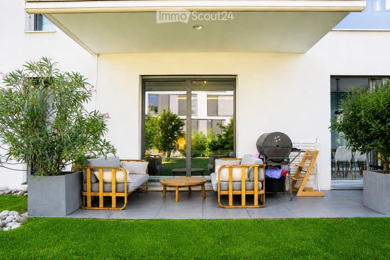 Modern outdoor patio with a sofa, chairs, a coffee table, and a barbecue, surrounded by greenery and a well-maintained lawn.