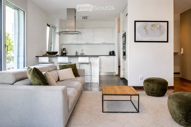 Modern living room with a grey sectional sofa, wooden coffee table, green ottomans, and a kitchen area with white cabinets, a kitchen island, and bar stools.