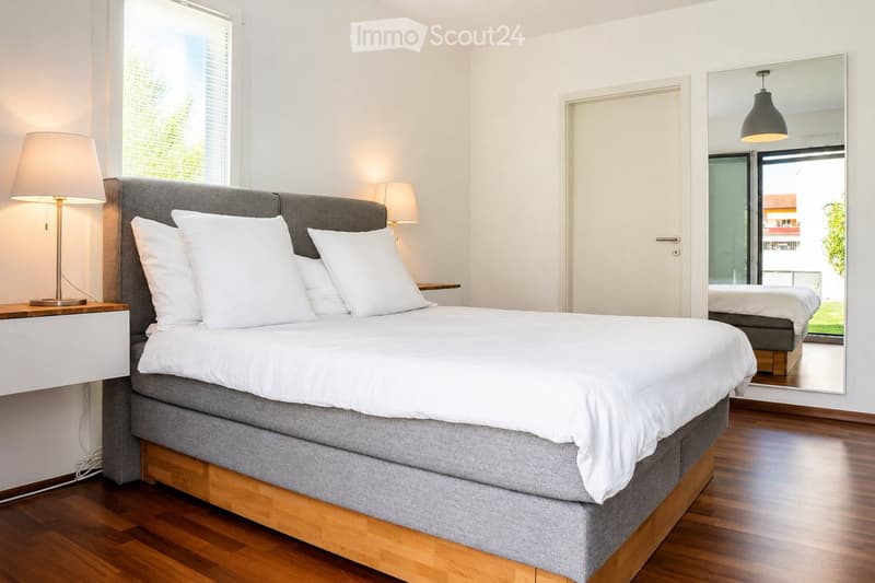 Modern bedroom with a large bed, white bedding, nightstand with lamp, and a large mirror reflecting part of the room.