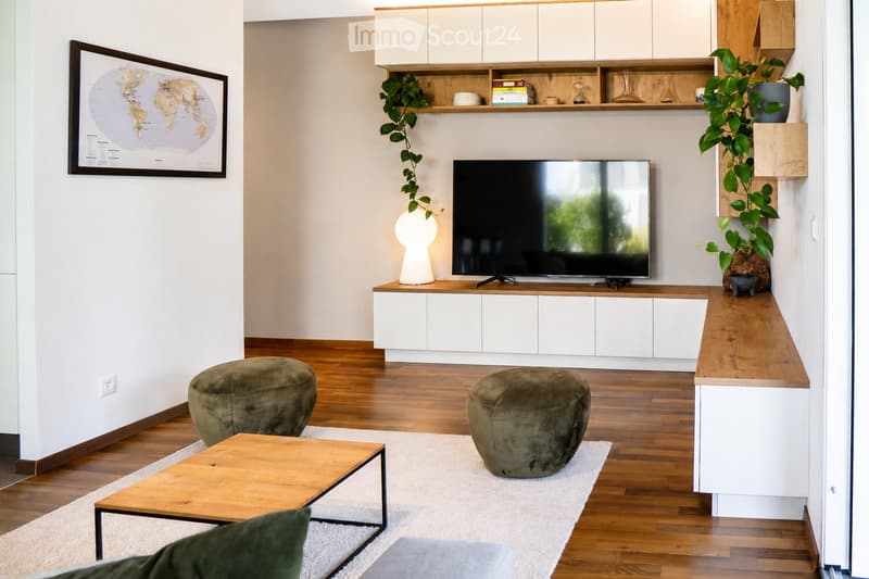 Modern living room with wooden flooring, white walls, and a neutral color palette. Features a television on a white cabinet, two green ottomans, a wooden coffee table, a floor lamp, and built-in shelving with decorative items and plants.