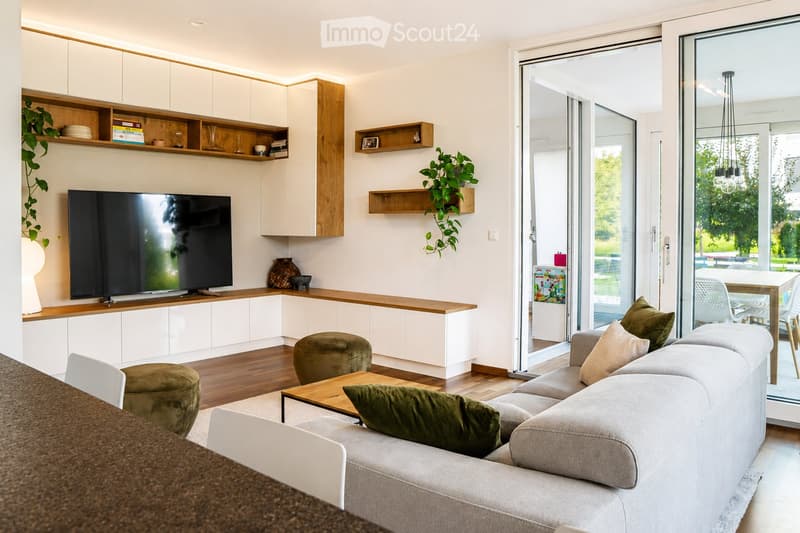 Modern living room with a grey sofa, wooden coffee table, green cushions, and a television on a white cabinet. The room features built-in white cabinets, wooden shelves, and a large glass sliding door leading to a patio.