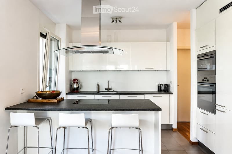 Modern kitchen with white cabinets, black countertops, and a central island with three white bar stools. Features include a built-in oven, a stand mixer, a coffee maker, and a range hood.