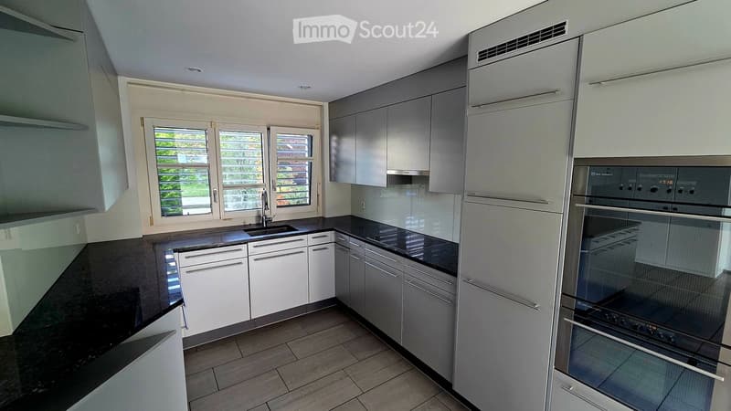 Modern kitchen with white and grey cabinets, black countertops, and a stainless steel oven. Features a large window with blinds and a sink with a faucet.