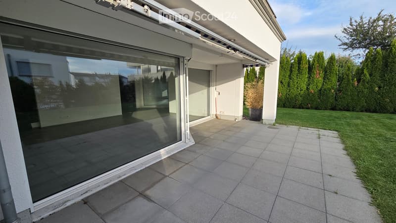Modern glass sliding door opens to a tiled balcony area with a view of the garden and surrounding trees.