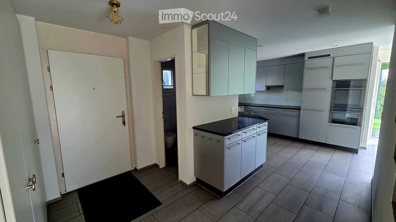 Modern kitchen with grey cabinets, black countertop, stainless steel appliances, and a built-in oven. The space includes a sink, a mirror, and a tiled floor.