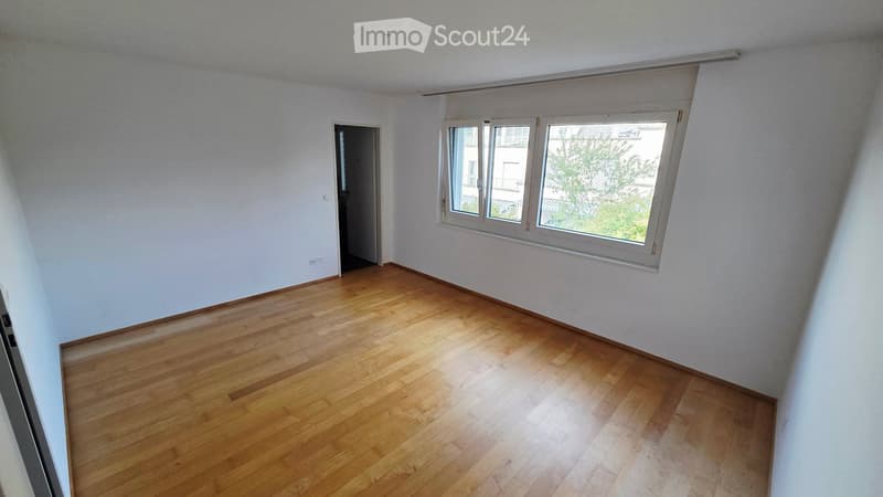 Empty room with wooden flooring and white walls, featuring a large window with blinds and a door leading to another space.