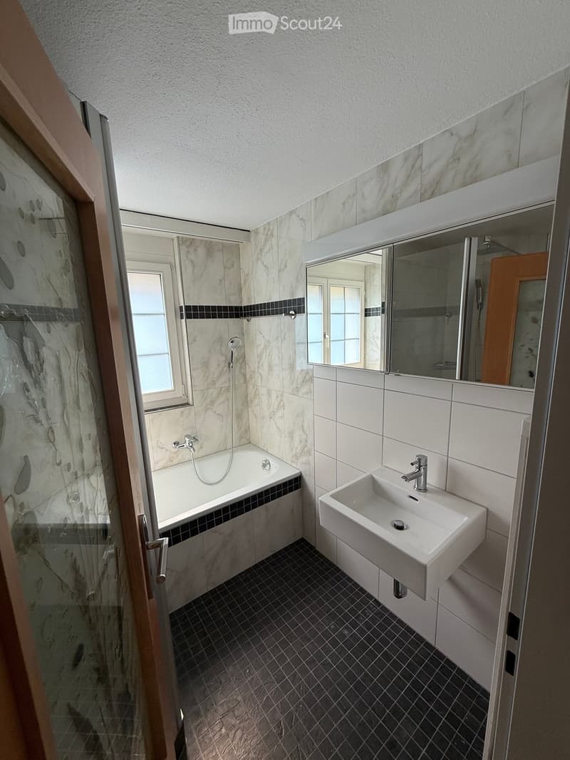 Modern bathroom with a bathtub, sink, and mirror. Features marble tiles on walls and dark tiled flooring.