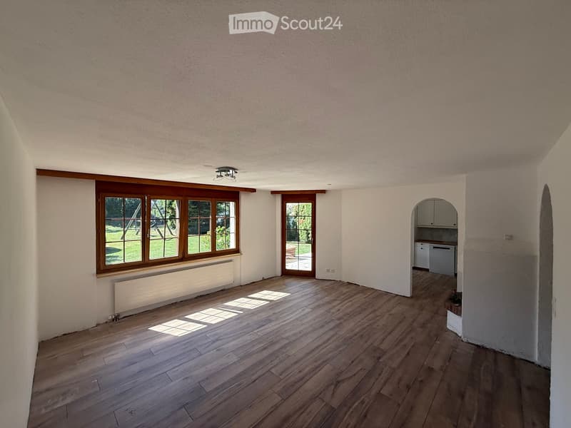 Empty room with wooden flooring, large windows, and a small kitchen area visible in the background.