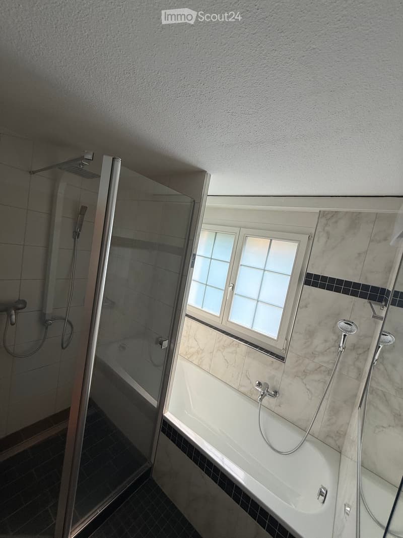Modern bathroom with a bathtub, glass-enclosed shower, and a window above the tub. The walls are tiled in marble, and the floor is dark tiled.