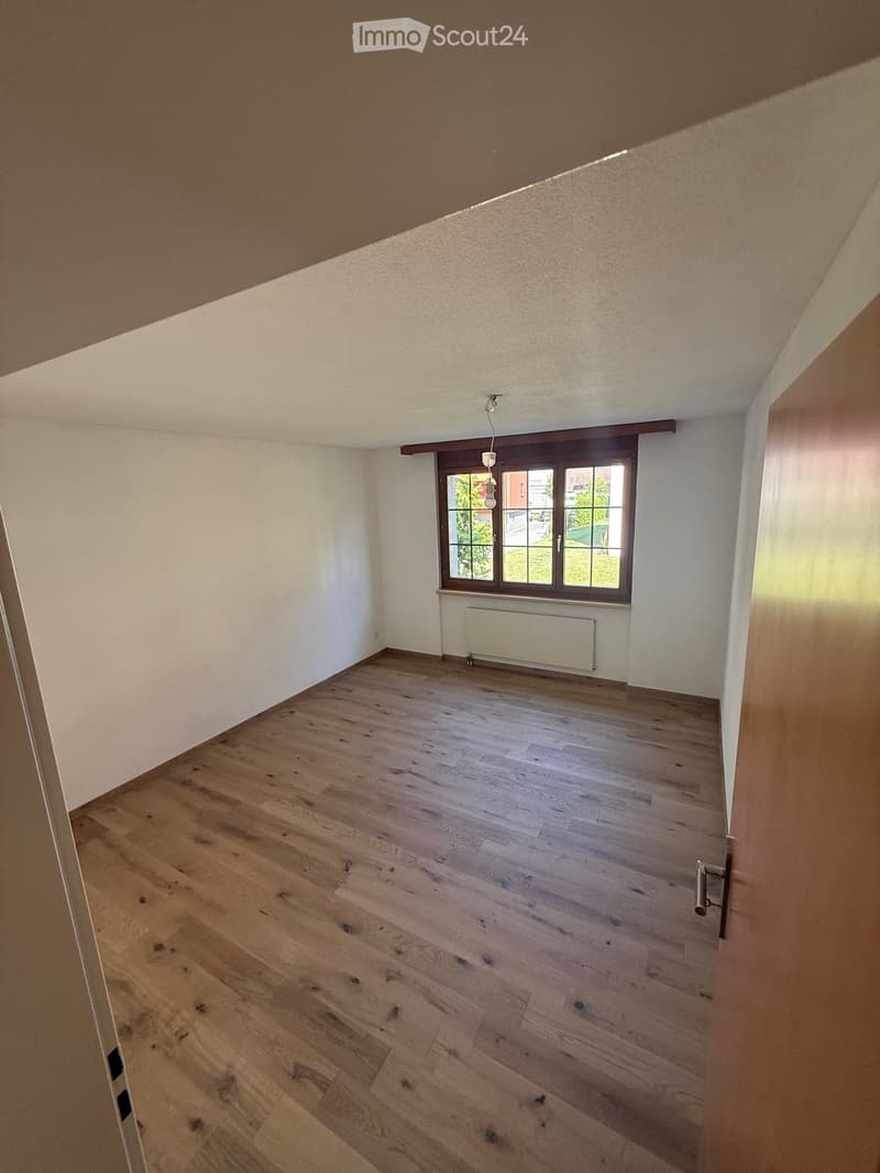 Empty room with wooden flooring, white walls, and a large window with brown frame. No furniture or equipment present.