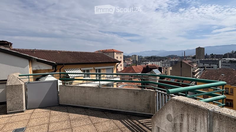 Balcony with green metal railing, overlooking a cityscape with buildings and distant mountains.