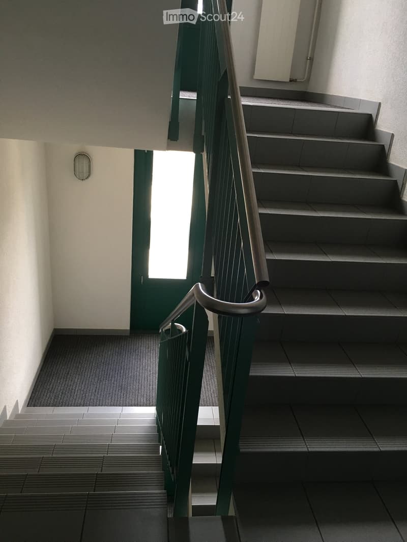 Staircase with green railing, gray tiled steps, and a small window at the top, leading to a hallway with light-colored walls and a small wall-mounted light fixture.
