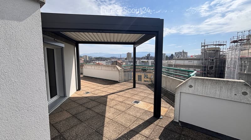 Balcony with tiled floor, metal railing, and covered section, overlooking a cityscape with buildings and mountains in the distance.