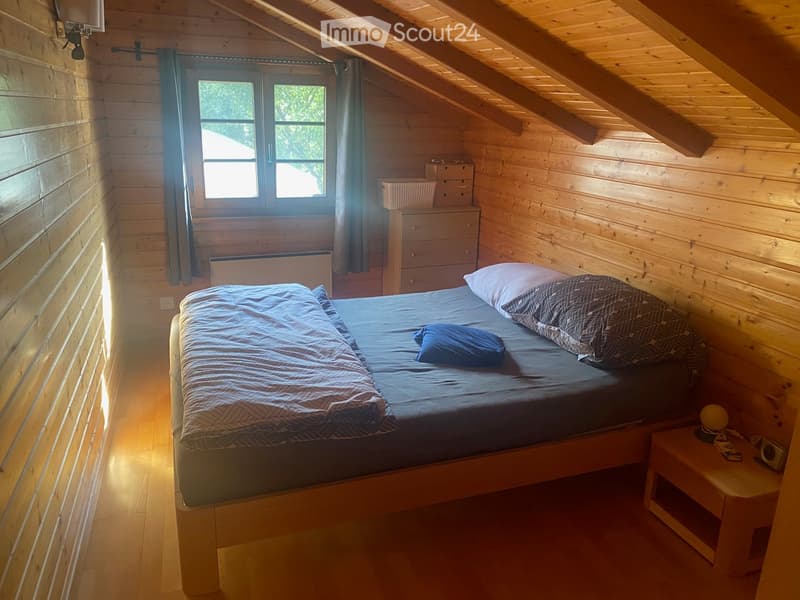 A small bedroom with wooden walls, a bed, a nightstand, a dresser, a radiator, a basket on top of the dresser, and a window with a curtain.