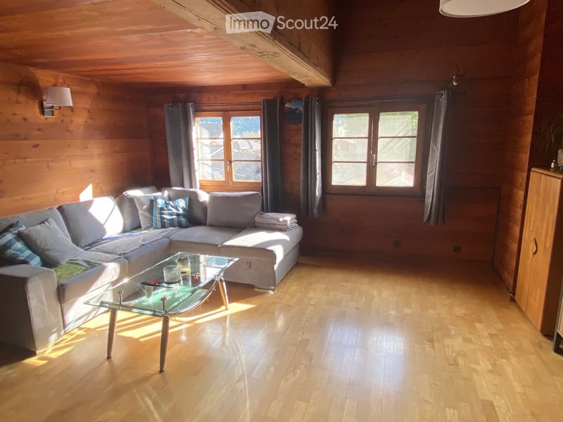 A cozy living room with wooden wall and floor, a grey sofa, glass coffee table, mounted lamps and large windows with gray curtains.