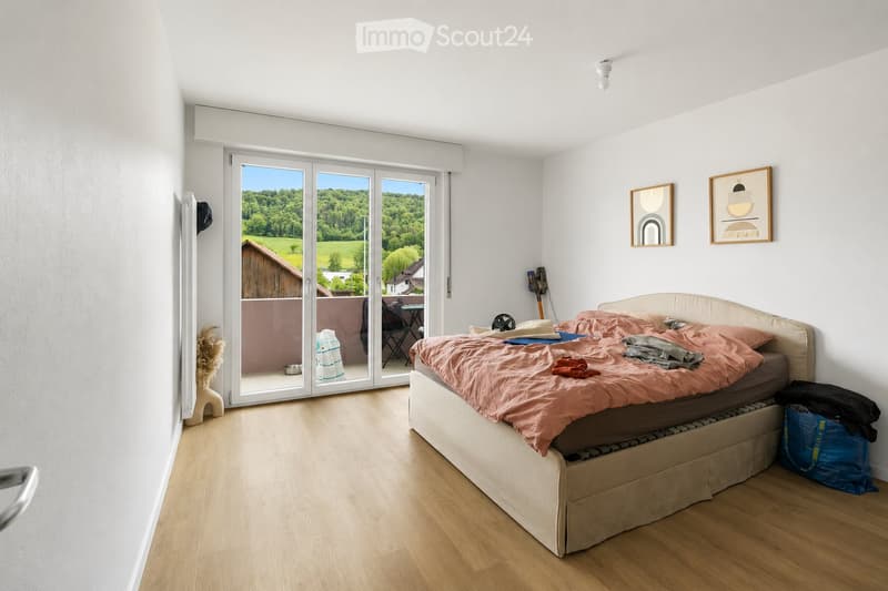 Bright bedroom with wooden flooring, a large bed with pink bedding, a sliding glass door leading to a balcony with outdoor furniture, and framed artwork on the walls.
