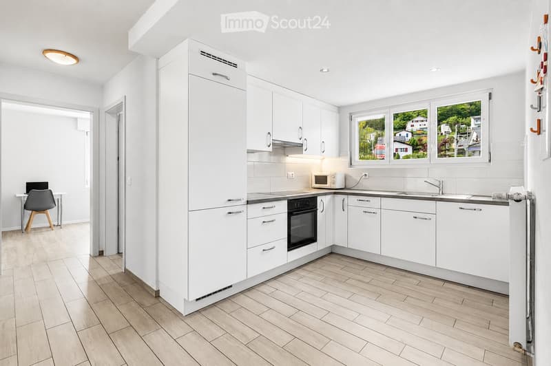 Modern kitchen with white cabinets, stainless steel appliances, and a wooden floor. Features a refrigerator, oven, microwave, sink, and a small workspace area with a chair and table.