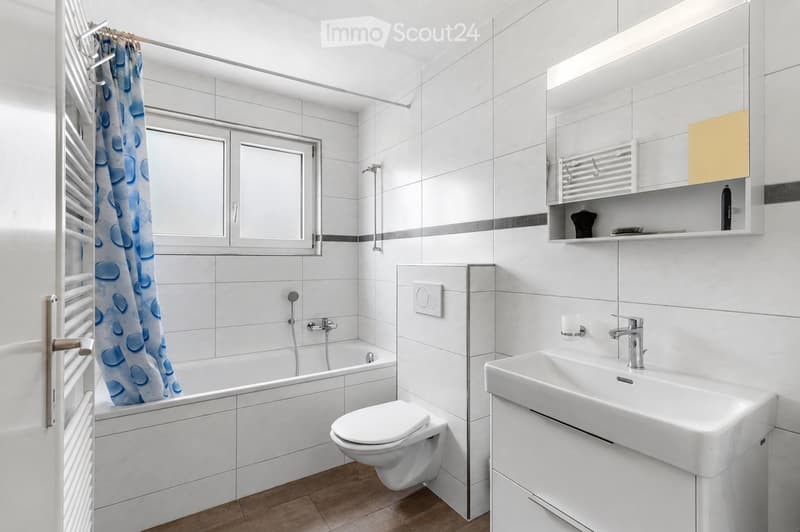 Modern bathroom with white tiles, a bathtub, toilet, sink, mirror, and a yellow cabinet. Features a window with a blue shower curtain and a radiator.