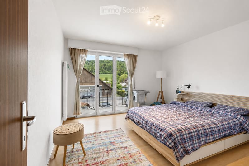 Bright bedroom with wooden floor, large sliding glass doors leading to a balcony, bed with plaid bedding, small round stool, floor lamp, and cat tree.