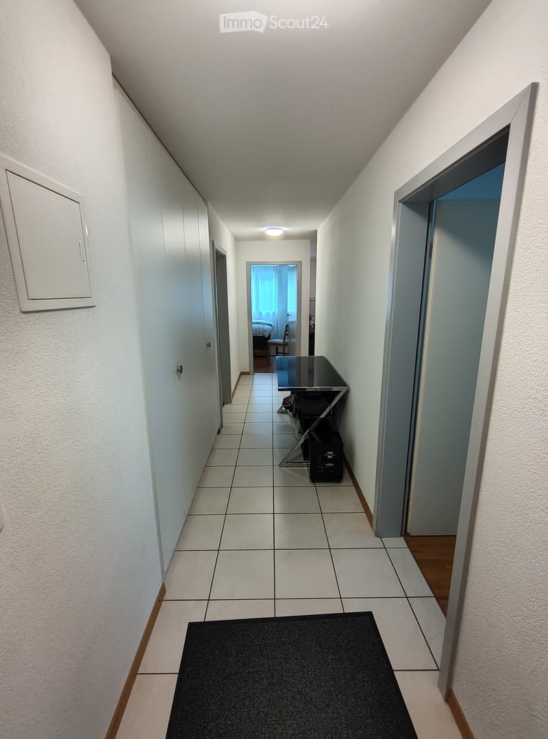 A narrow hallway with white walls, tiled floor, and a black doormat at the entrance. A small table and chair are visible in the background, along with a glimpse of a bedroom.
