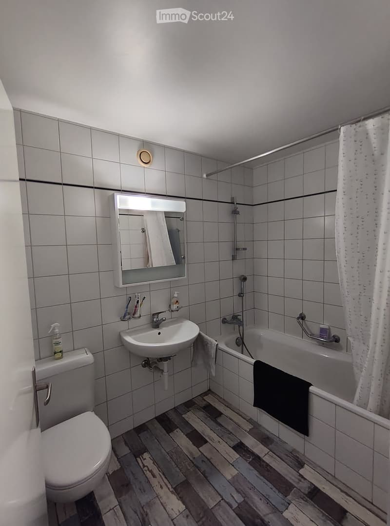 Modern bathroom with wooden flooring, white tiled walls, a bathtub with a black towel, a sink with a mirror cabinet, and a toilet.