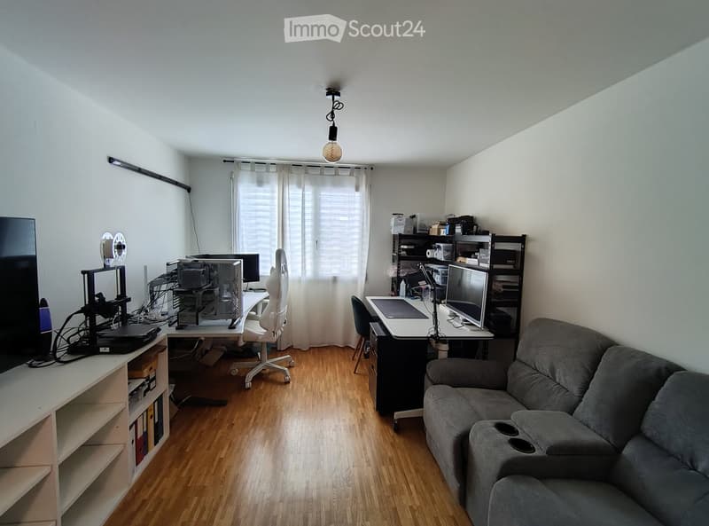 A multi-functional room with a sofa, desk, and computer setup, featuring wooden flooring and large windows with curtains.
