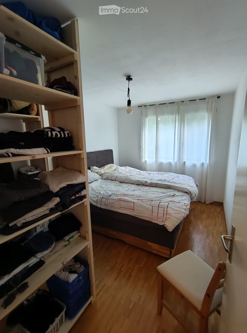 A small bedroom with a single bed, a wooden chair, and a tall shelf filled with folded clothes and storage boxes. The room has wooden flooring and white walls.